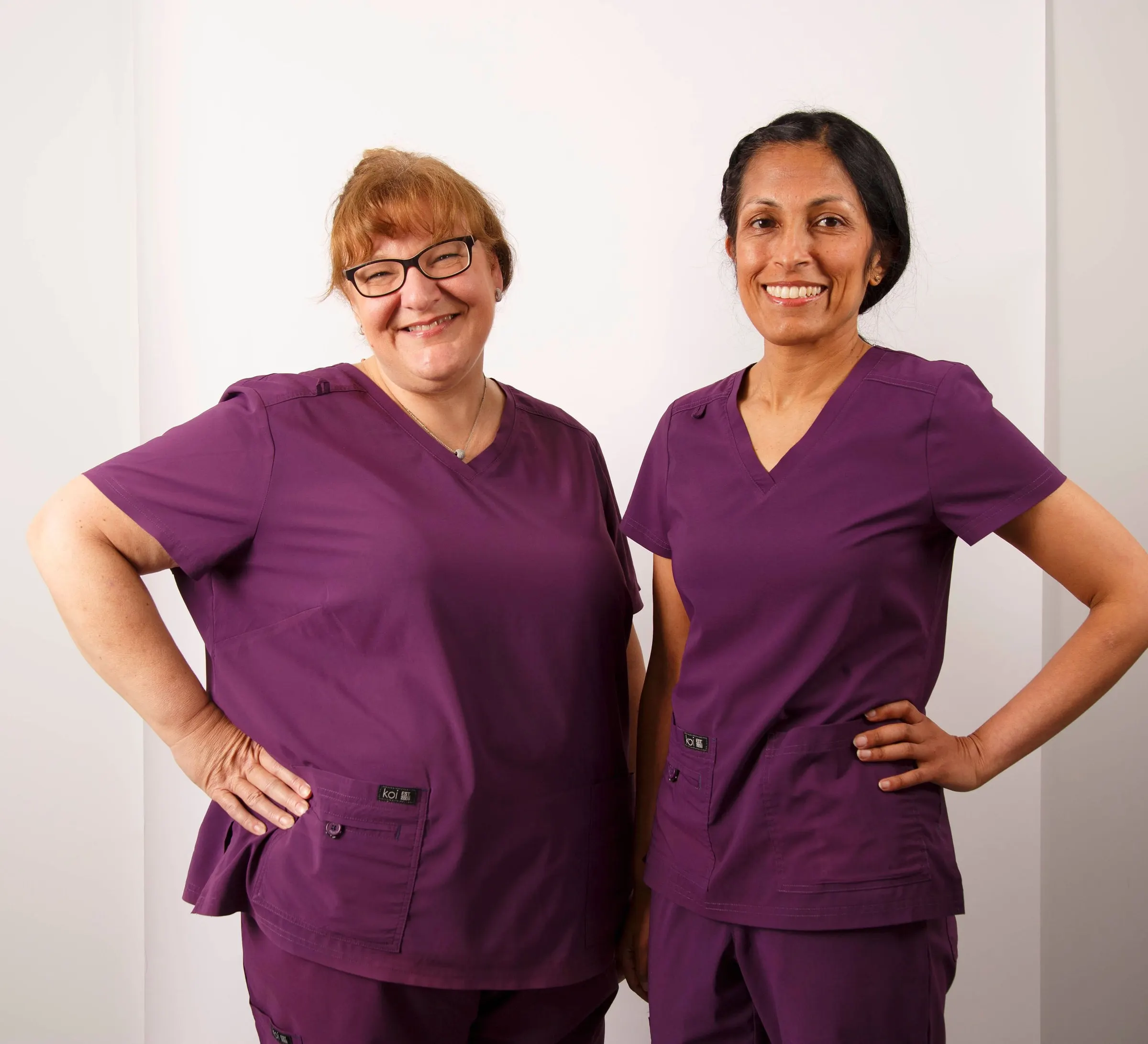 Consultant orthodontists Anne-Marie Smith and Anjli Patel in purple scrubs, smiling with hands on hips